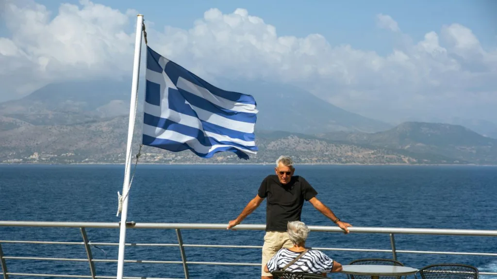 die griechische Flagge auf einem Schiff weht im Wind und ein Rentnerpaar ist an der Reeling