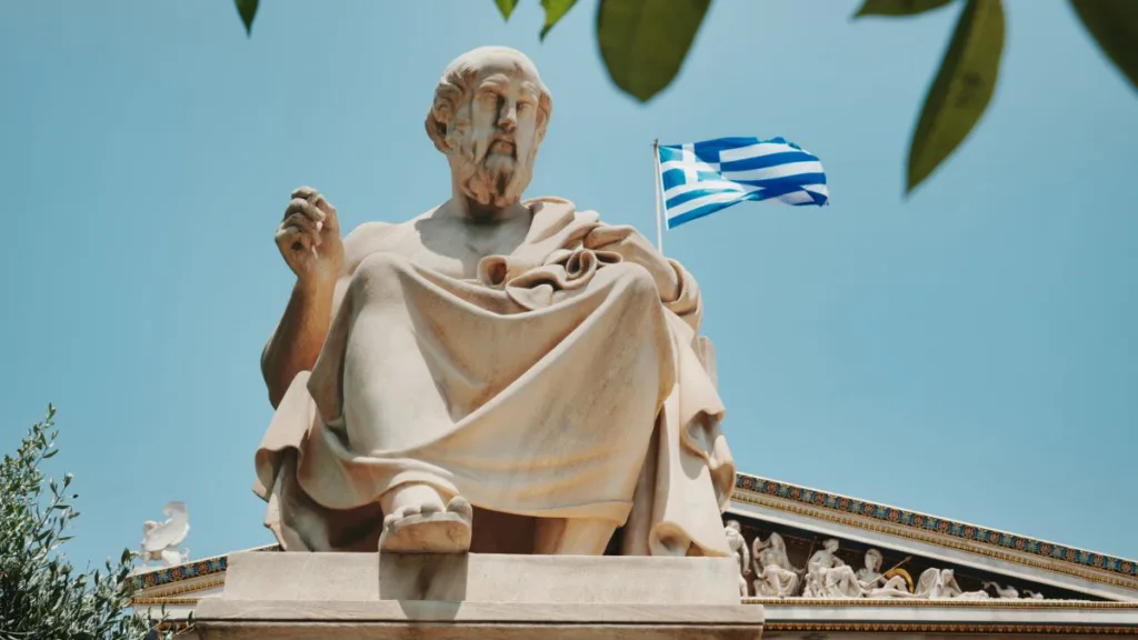 Statue eines griechischen Philosophen vor blauem Himmel mit wehender Griechenland-Flagge im Hintergrund.