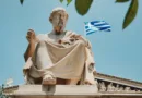 Statue eines griechischen Philosophen vor blauem Himmel mit wehender Griechenland-Flagge im Hintergrund.