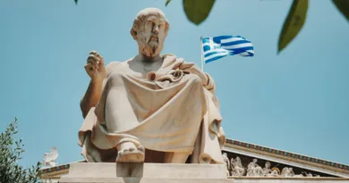 Statue eines griechischen Philosophen vor blauem Himmel mit wehender Griechenland-Flagge im Hintergrund.