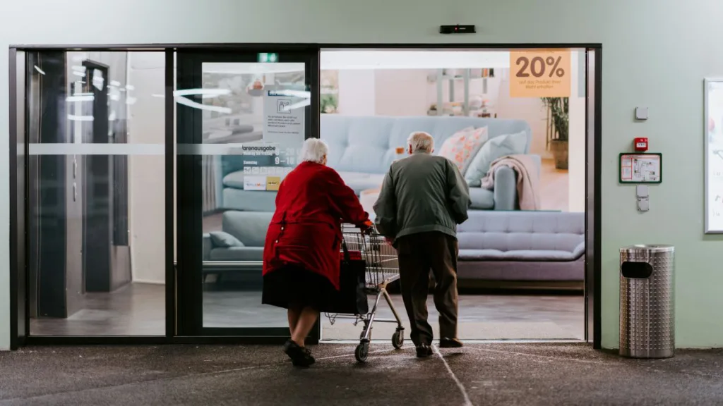 Zwei ältere Menschen gehen vor einer hell beleuchteten Schaufensterfront in einen Laden.