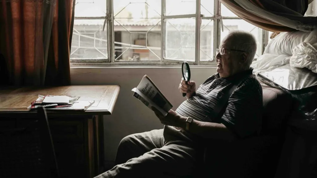 ein Senior liest am Fenster eine Zeitung mit einer Leselupe